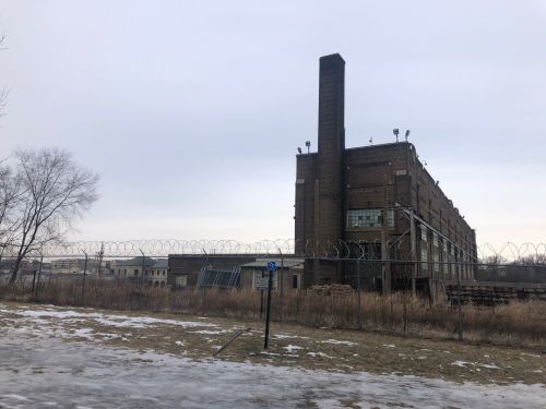 An old industrial building with a tall chimney, surrounded by a barbed wire fence and a grassy area, under a cloudy sky.