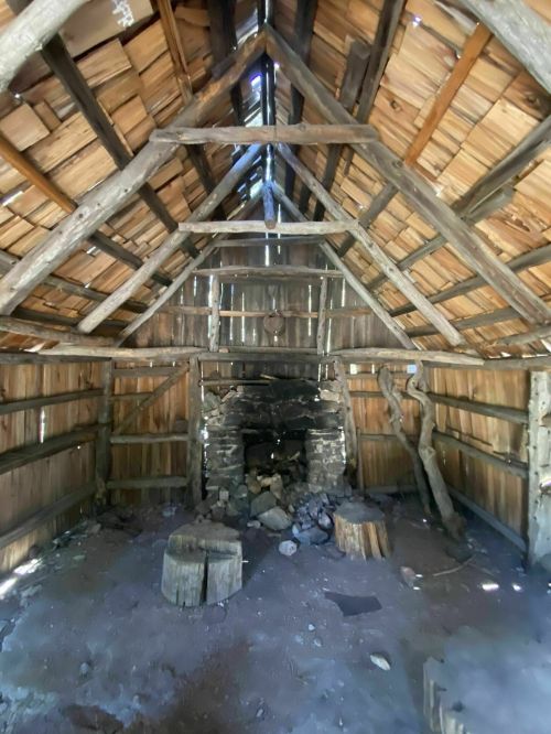 Interior of a rustic cabin with wooden beams, a stone fireplace, and a dirt floor, illuminated by natural light.