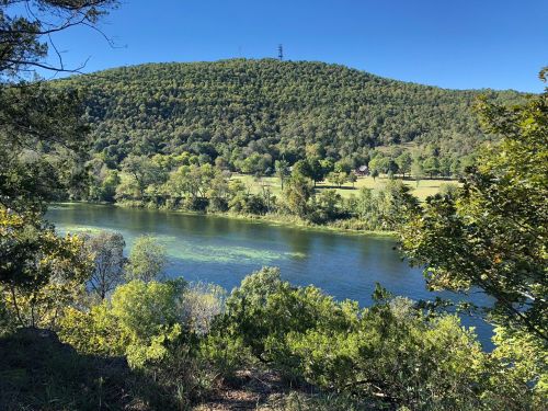 A serene river flows beside a lush green hillside under a clear blue sky. Trees frame the peaceful landscape.