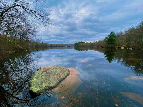 A calm lake reflects a cloudy sky, surrounded by trees and a large rock in the foreground.