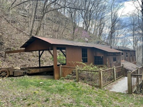 A rustic wooden building with a metal roof, surrounded by trees and a path, set in a hilly landscape.