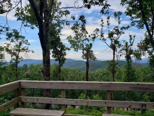 A wooden overlook with a view of lush green mountains and a blue sky dotted with clouds.