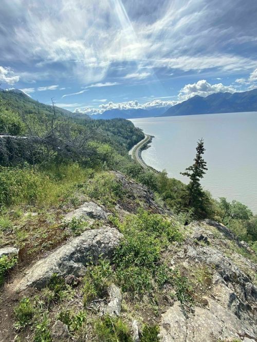 A scenic view of a winding road along a river, surrounded by lush greenery and mountains under a partly cloudy sky.