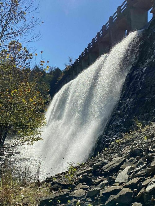 A waterfall cascading over a stone dam, surrounded by trees and a clear blue sky.