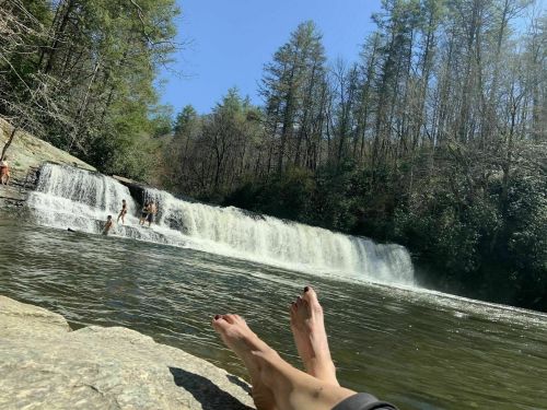 A serene waterfall surrounded by trees, with people swimming and a pair of feet resting on a rock in the foreground.