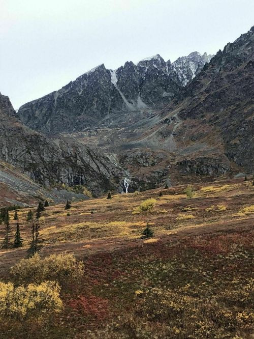 A scenic mountain landscape with rugged peaks, colorful autumn foliage, and a hint of a waterfall in the distance.