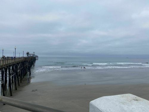 A cloudy beach scene featuring a pier, gentle waves, and a few surfers in the water.