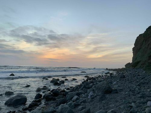 A rocky beach at sunset, with gentle waves and a cliff in the background under a colorful sky.
