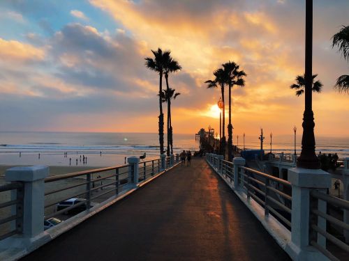 A sunset view over a pier lined with palm trees, with people walking along the path and waves in the background.