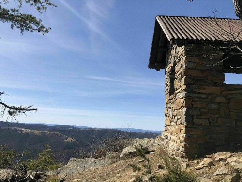 A stone structure with a metal roof overlooks a vast mountainous landscape under a clear blue sky.