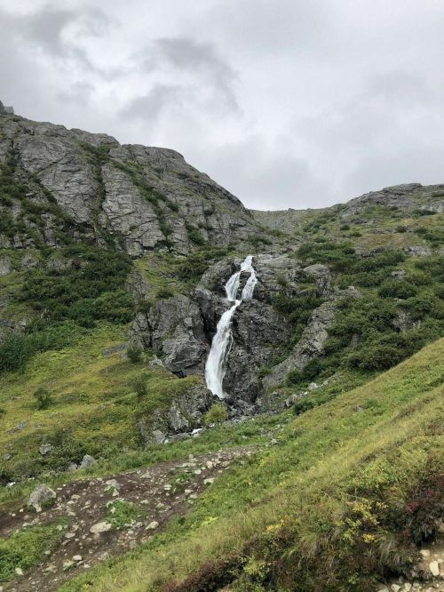 A waterfall cascades down rocky terrain, surrounded by lush greenery under a cloudy sky.