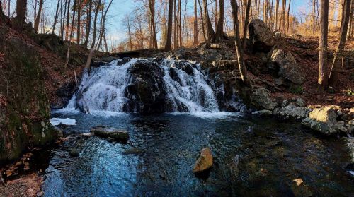 A serene waterfall cascades over rocks into a clear pool, surrounded by trees and autumn foliage.