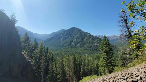 A panoramic view of lush green mountains under a clear blue sky, with trees in the foreground and distant peaks.