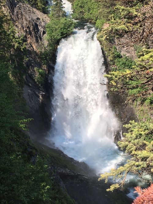 A cascading waterfall surrounded by lush greenery and rocky terrain, with mist rising from the water.
