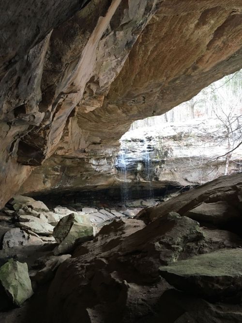 A rocky cave interior with a small waterfall and sunlight filtering through, surrounded by natural stone formations.