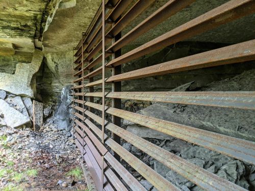 Rusty metal bars cover an opening in a rocky cave, revealing dark stone and scattered debris inside.