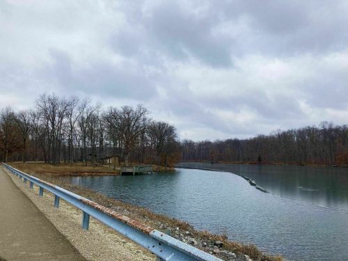 A serene lake surrounded by bare trees under a cloudy sky, with a path and a small dock visible along the shore.