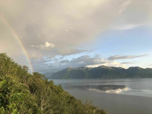 A serene landscape featuring mountains, a calm lake, and a rainbow arching across a cloudy sky.