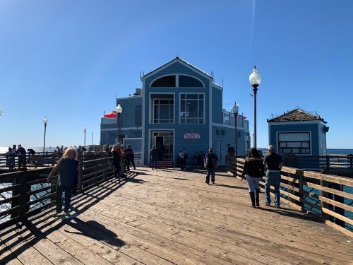 A busy pier with a blue building at the end, people walking, and clear skies above.