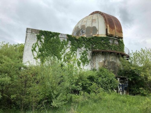 Abandoned observatory covered in green vines, with a rusty dome, surrounded by overgrown vegetation under a cloudy sky.