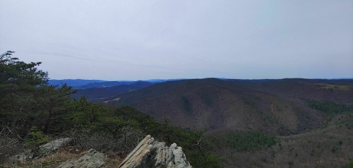 A panoramic view of rolling mountains under a cloudy sky, with sparse trees and rocky foreground.