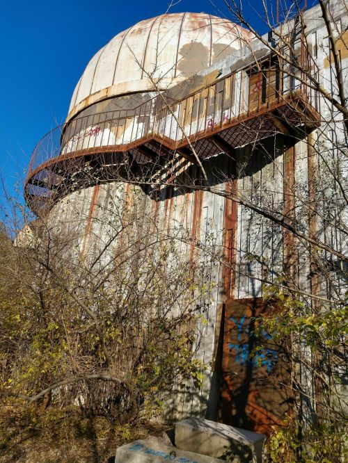 Abandoned building with a dome roof, rusted exterior, and overgrown vegetation surrounding it under a clear blue sky.