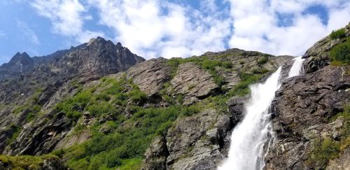 A cascading waterfall flows down rocky terrain, surrounded by lush greenery and under a blue sky with scattered clouds.