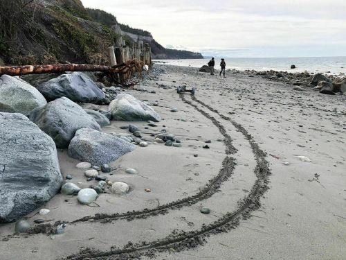 A sandy beach with large rocks and tire tracks leading towards two people walking in the distance.