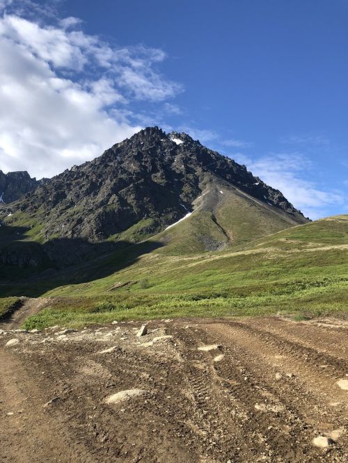 A rugged mountain peak rises against a blue sky, surrounded by green hills and a dirt path in the foreground.
