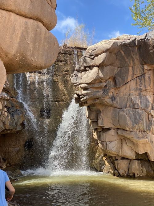 A waterfall cascades between large rock formations, creating a serene natural scene under a blue sky.