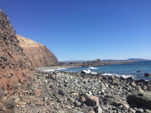 Rocky shoreline with waves crashing against the stones, under a clear blue sky. Coastal cliffs in the background.