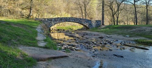 A stone arch bridge spans a shallow stream, surrounded by trees and grassy banks in a serene natural setting.