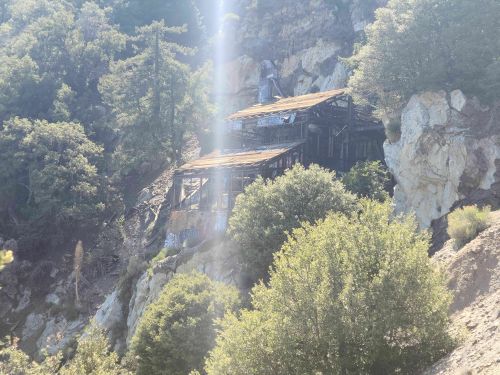 Abandoned structure on a rocky hillside, surrounded by trees and overgrown vegetation, with graffiti on the walls.