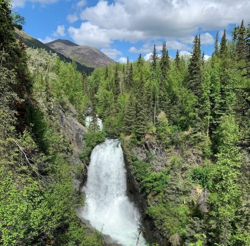 A cascading waterfall surrounded by lush green trees and mountains under a partly cloudy sky.
