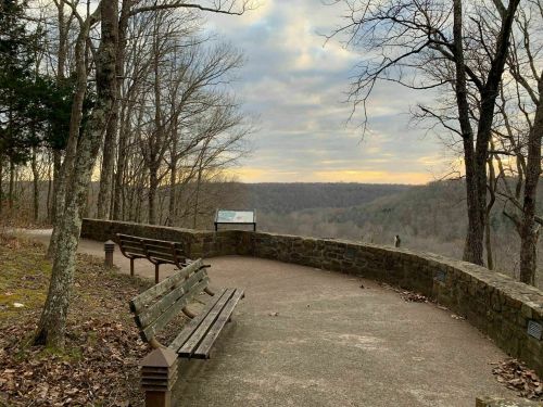 A scenic overlook with benches, surrounded by trees, offering a view of rolling hills under a cloudy sky.