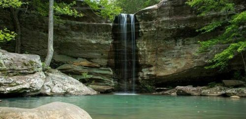 A serene waterfall cascades into a clear pool, surrounded by lush greenery and rocky cliffs.