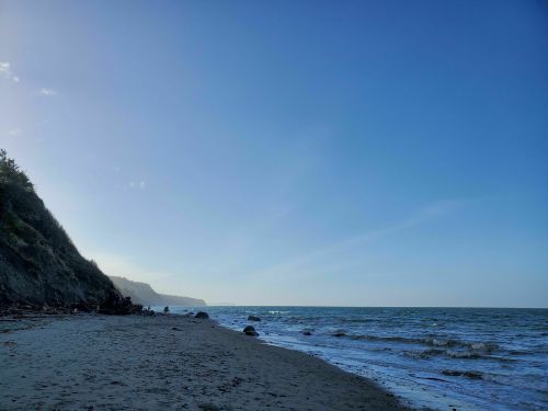 A serene beach scene with gentle waves, a clear blue sky, and a distant cliff in the background.