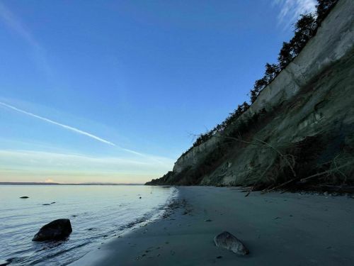 A serene beach scene with a calm shoreline, cliffs in the background, and a clear blue sky.