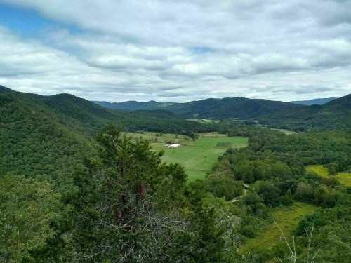 A panoramic view of rolling green hills and fields under a cloudy sky, surrounded by dense forests.