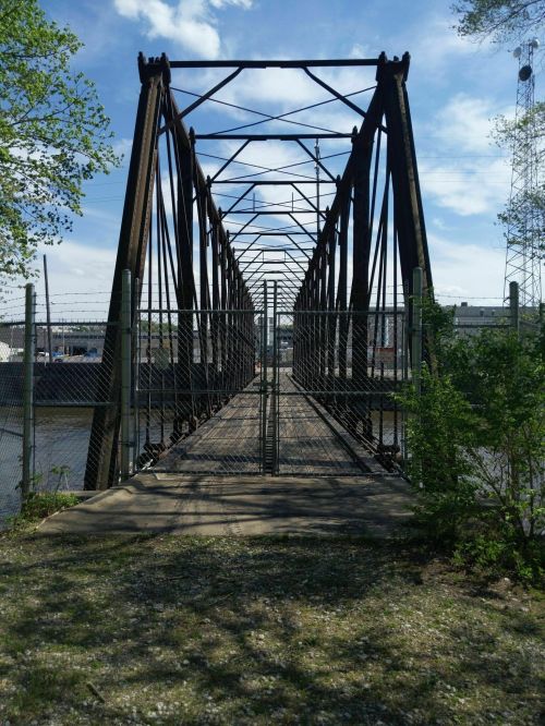 A metal truss bridge spans a river, surrounded by greenery and a fence, under a blue sky with scattered clouds.