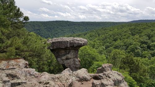 A rocky outcrop with a flat top overlooks a lush green forest and rolling hills under a cloudy sky.