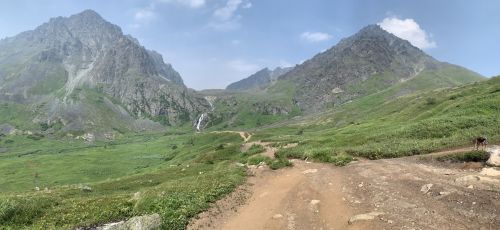A scenic mountain landscape with a dirt path, lush greenery, and a waterfall cascading down rocky cliffs.