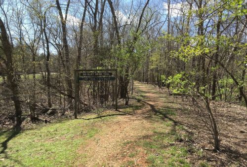 A dirt path leads into a wooded area, marked by a sign for the Bob Elliott Nature Trail. Trees and greenery surround the trail.