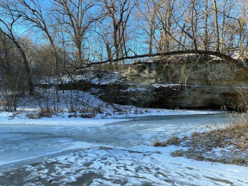 A snowy landscape with a frozen river, rocky cliffs, and bare trees under a clear blue sky.