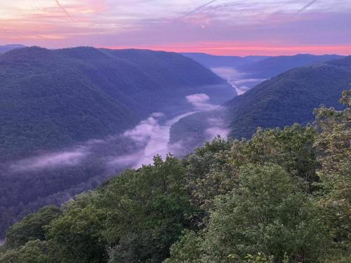A serene view of a river winding through lush green mountains at sunrise, with mist rising from the water.