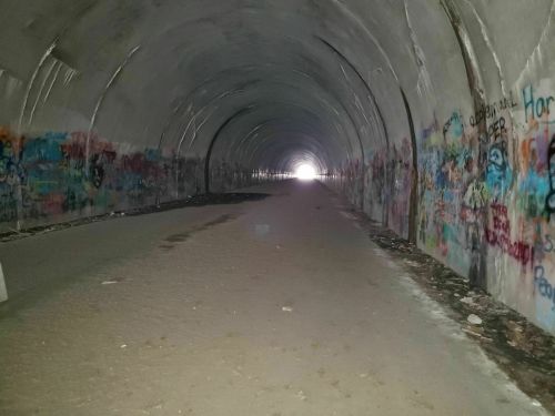 A dark tunnel with graffiti-covered walls, leading to a bright light at the far end. The ground is sandy and uneven.