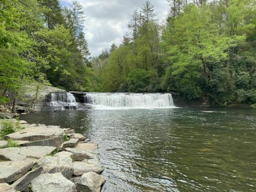 A serene waterfall cascades into a calm river, surrounded by lush green trees and rocky shoreline.