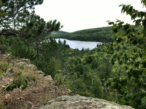 A scenic view of a lake surrounded by lush green trees and hills, seen from a rocky vantage point.