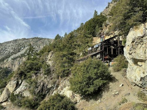 Abandoned mining structure on a rocky hillside, surrounded by trees and mountains under a blue sky.