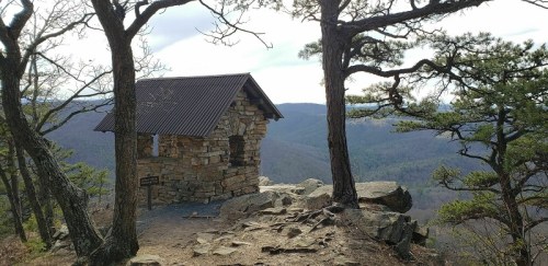 A small stone cabin on a rocky outcrop, surrounded by trees and overlooking a mountainous landscape.